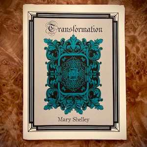 Puede incluir: Una portada de libro con un marco blanco y negro y un diseño ornamentado en azul turquesa y negro. El título "Transformation" está escrito en una fuente decorativa. El autor "Mary Shelley" está escrito debajo del diseño.