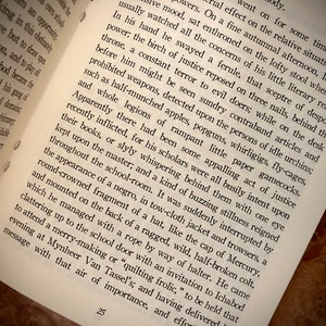 K&ouml;nnte beinhalten: Offenes Buch mit Text auf einer Holzoberfl&auml;che. Die Seiten sind leicht vergilbt und der Text ist schwarz. Das Buch ist auf einer Seite mit der Nummer 25 unten ge&ouml;ffnet. Der Text beschreibt eine Szene mit einem Schullehrer und Sch&uuml;lern.