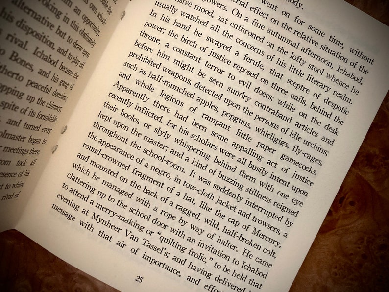 K&ouml;nnte beinhalten: Offenes Buch mit Text auf einer Holzoberfl&auml;che. Die Seiten sind leicht vergilbt und der Text ist schwarz. Das Buch ist auf einer Seite mit der Nummer 25 unten ge&ouml;ffnet. Der Text beschreibt eine Szene mit einem Schullehrer und Sch&uuml;lern.