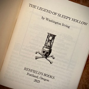 K&ouml;nnte beinhalten: Aufgeschlagenes Buch mit dem Titel "THE LEGEND OF SLEEPY HOLLOW" von Washington Irving. Eine Schwarz-Wei&szlig;-Illustration eines Sch&auml;dels mit einer Sanduhr und gekreuzten Knochen befindet sich auf der Seite. Das Buch stammt von Renfield's Books, Portland, Oregon, 2023.
