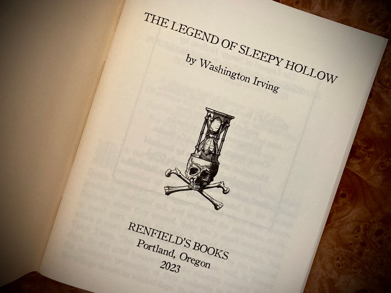 K&ouml;nnte beinhalten: Aufgeschlagenes Buch mit dem Titel "THE LEGEND OF SLEEPY HOLLOW" von Washington Irving. Eine Schwarz-Wei&szlig;-Illustration eines Sch&auml;dels mit einer Sanduhr und gekreuzten Knochen befindet sich auf der Seite. Das Buch stammt von Renfield's Books, Portland, Oregon, 2023.