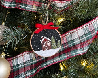 May include: A hand-painted wooden ornament featuring a small house with a snowy roof and two pine trees. The ornament is painted on a round piece of wood with a black background and white snow. The house is brown with white trim and a red roof. The ornament is tied with a red ribbon.
