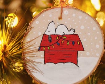 May include: A wooden Christmas ornament with a hand-painted design of Snoopy sleeping on top of his red doghouse with a string of colorful lights. The ornament is decorated with white paint to resemble snow.