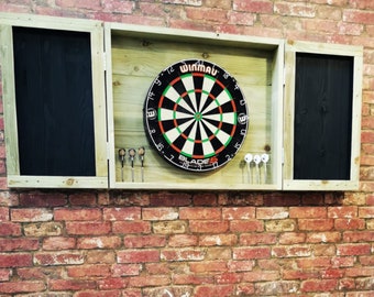 Op de afbeelding: Een houten dartbordkast met twee deuren, één open en één gesloten. De kast heeft een zwart schoolbord op elke deur. De kast is gemonteerd op een bakstenen muur. Een dartbord is zichtbaar in het midden van de kast. Het dartbord is zwart en groen met een rode bullseye. Het dartbord is gelabeld "Winmau Blade 5".
