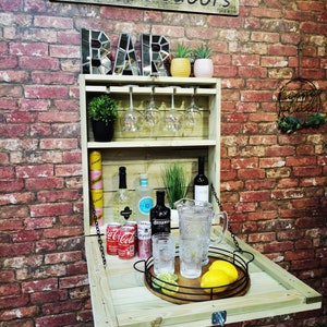 May include: A wooden outdoor bar with the text "Bars Outdoors" above. The bar has a shelf for glasses, bottles, and a serving tray with lemons and limes. The bar is attached to a red brick wall.
