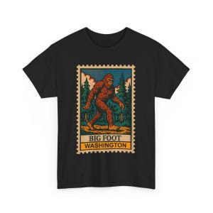 Puede incluir: Camiseta negra con un diseño de sello de Bigfoot. La ilustración del sello muestra un Bigfoot marrón caminando en un bosque con árboles verdes y un cielo azul. El sello dice "BIG FOOT WASHINGTON" en una pancarta amarilla y blanca.