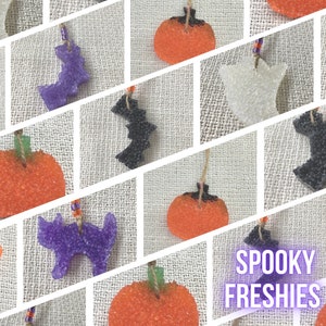 May include: A collection of Halloween-themed air fresheners in various shapes, including pumpkins, bats, and ghosts. The air fresheners are made of a textured, bubbly material and come in a variety of colors, including orange, purple, black, and white. The text "SPOOKY FRESHIES" is written in purple letters on a white background.