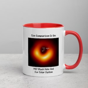 以下が含まれることがあります： 赤色の内側とハンドルが付いた白いセラミックマグ。マグカップには、テキスト「Size Comparison Of the M87 Black Hole And Our Solar System」とともに、M87ブラックホールのグラフィックが描かれています。