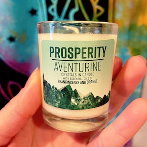 May include: A small glass candle labeled "PROSPERITY AVENTURINE" with text indicating crystals and essential oils of frankincense and orange. The candle has a light green label with a green crystal design at the bottom. The candle is held in a hand.