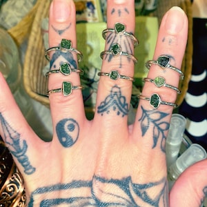 May include: A collection of silver rings, each featuring a raw, green gemstone. The rings are displayed on fingers with intricate tattoos. The gemstones vary in shape and size, set in simple silver bands.