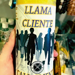May include: Yellow candle in a glass jar with the words "LLAMA CLIENTE" and "CALL CLIENTS" printed on the label. The label also features a silhouette of people. The candle is from the Lázaro Brand.