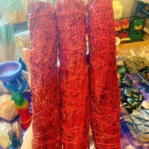 May include: Three bundles of red sage, tightly wrapped with red twine. The sage has a textured, fibrous appearance. The bundles are held in a person's hand, with other spiritual items visible in the background.