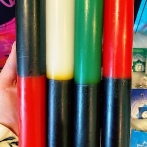 May include: Four pillar candles in a row, each with a different color scheme. The top halves are black, white, green, and red, respectively. The bottom halves are black. The candles are cylindrical and appear to be made of wax.