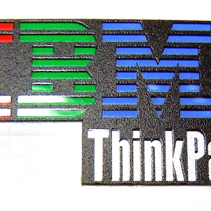 May include: A black IBM ThinkPad logo with red, green, and blue detailing. The logo is on a textured surface, with the words "ThinkPad" in white text below the IBM logo.