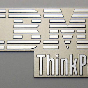 May include: A silver IBM ThinkPad logo on a beige background. The logo is made up of a series of lines that form the letters IBM. The words ThinkPad are below the IBM logo.