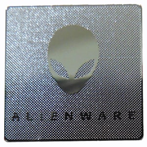 Könnte beinhalten: Quadratische, silberfarbene Metallplatte mit strukturierter Oberfläche. Sie zeigt einen Ausschnitt eines Alienkopfes in der Mitte und das Wort "ALIENWARE" am unteren Rand. Das Design ist schlicht und modern.