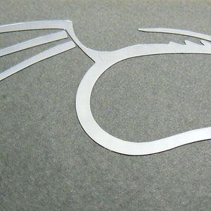 Peut inclure: Une découpe en métal argenté d'un dragon stylisé avec des ailes déployées. Le dragon est tourné vers la droite et a une longue queue recourbée.