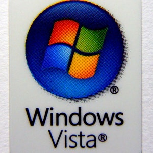 Könnte beinhalten: Ein weißer Aufkleber mit einem blauen Kreis, der das Windows Vista-Logo enthält. Das Logo ist ein vierflügeliges Fenster in den Farben Blau, Grün, Orange und Rot. Der Text "Windows Vista" befindet sich unter dem Logo.