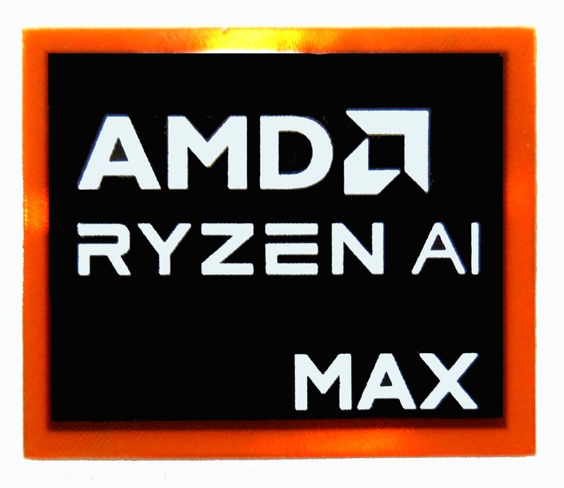 May include: A rectangular sticker with an orange border and a black center. The white text reads "AMD RYZEN AI MAX". The AMD logo is also white. The sticker is likely for a computer component.