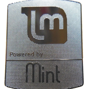 Puede incluir: Una insignia rectangular plateada con una superficie texturizada. La insignia presenta el logotipo estilizado "LM" y el texto "Powered by Mint" y "Mint" en color oscuro. El diseño general es simple y moderno.