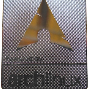 May include: A silver-colored, rectangular metal badge with a textured surface. The badge features a gold-colored, stylized arch logo at the top and the words "Powered by" and "archlinux" in black text.