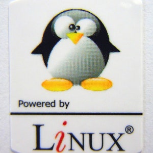 May include: A white sticker with a cartoon penguin in black, gray, and yellow. The penguin is wearing a black tuxedo and has a yellow beak. The text "Powered by Linux" is printed below the penguin.