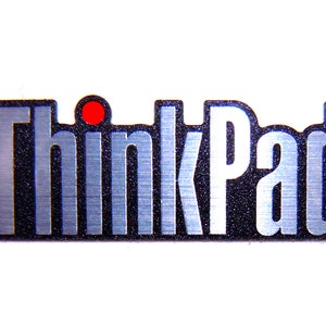 May include: A silver ThinkPad logo with a red dot above the "i" in Think. The logo is on a black background with a brushed metal finish.