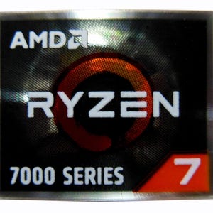 Peut inclure: Autocollant rectangulaire avec le logo AMD en haut. Le mot "RYZEN" est en blanc, avec un design circulaire rouge et orange en arrière-plan. Le texte "7000 SERIES" est en dessous, avec le chiffre "7" dans un coin rouge.