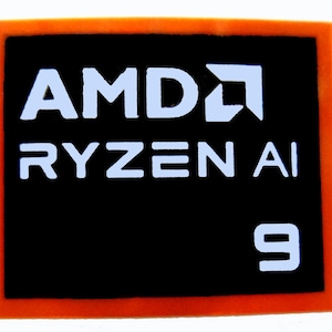 May include: A rectangular sticker with an orange border and a black centre. The white text reads "AMD RYZEN AI 9". The AMD logo is also in white.