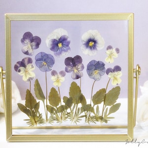May include: A gold-framed picture with a pressed flower arrangement of purple and white pansies. The flowers are arranged in a row with their stems and leaves visible. The picture is on a white surface.