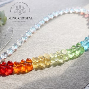 Puede incluir: Correa para teléfono con un dije de osito gomoso arcoíris y una cadena de cristal transparente. El dije presenta un arcoíris de colores, desde el rojo hasta el morado. La correa está unida a un clip plateado. "Bring Sparkle to Everyday Life" y "Bling Crystal Handmade Jewellery" están impresos en el dije.
