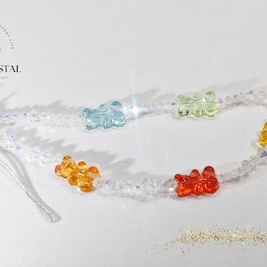 Puede incluir: Un collar con un dije de osito de gomita arcoíris y cuentas de cristal transparente. El collar tiene un cierre plateado y una cuerda para sujetarlo a un teléfono o bolso.