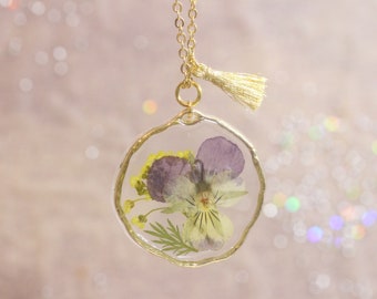 May include: A gold chain necklace with a round pendant featuring a pressed purple and yellow flower encased in clear resin. The pendant has a gold rim and a gold tassel hangs from the chain.