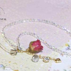 May include: A delicate phone charm featuring a preserved pink rose, clear crystal beads, and gold accents. The charm is attached to a thin, white string.