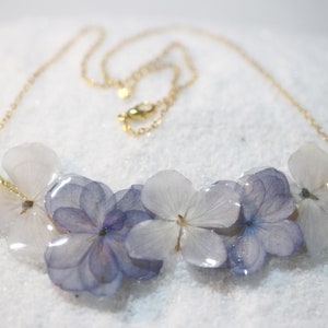 May include: A delicate gold chain necklace featuring a cluster of preserved hydrangea flowers encased in clear resin. The flowers are a pale lavender colour.