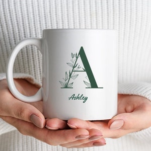 Può includere: Tazza in ceramica bianca con un design a foglie verdi e la lettera "A" al centro. Il nome "Ashley" è stampato sotto la lettera.