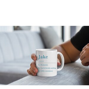 K&ouml;nnte beinhalten: Eine wei&szlig;e Keramiktasse mit blauem Design. Die Tasse sagt "Jake [Substantiv] 1. Workaholic. 2. Kann buchst&auml;blich alles reparieren. 3. Kaffeekenner."
