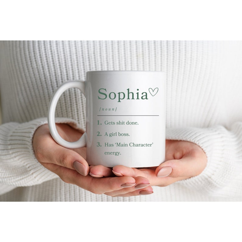 K&ouml;nnte beinhalten: Eine wei&szlig;e Keramiktasse mit gr&uuml;nem Design. Die Tasse tr&auml;gt die Aufschrift "Sophia" mit einem Herzsymbol und einer Definition des Namens: "Gets shit done. A girl boss. Has 'Main Character' energy."