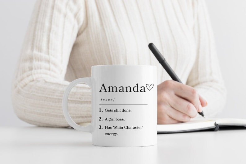 K&ouml;nnte beinhalten: Eine wei&szlig;e Keramiktasse mit einer schwarzen Aufschrift, die lautet "Amanda [Substantiv] 1. Macht den Job. 2. Eine Chefin. 3. Hat 'Hauptfigur'-Energie." Die Tasse hat ein schwarzes Herzsymbol neben dem Namen.