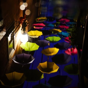 May include: A night scene with colorful umbrellas hanging overhead in a narrow alleyway. The umbrellas are yellow, blue, red, and green, and they are illuminated by streetlights. The alleyway is dark and wet, and the reflections of the umbrellas can be seen in the puddles on the ground.