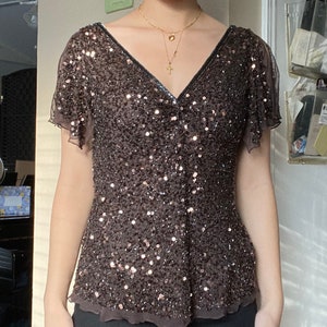 Puede incluir: Una blusa marr&oacute;n con lentejuelas, cuello en V y mangas cortas. La blusa tiene un dobladillo con volantes y est&aacute; hecha de una tela transparente.