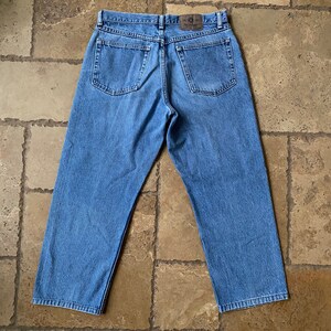 K&ouml;nnte beinhalten: Eine blaue Jeans aus Denim mit einem braunen Leder-Patch am hinteren Bund. Die Jeans hat eine lockere Passform und ein gerades Bein.