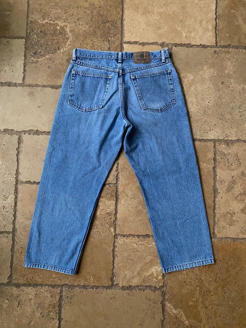 K&ouml;nnte beinhalten: Eine blaue Jeans aus Denim mit einem braunen Leder-Patch am hinteren Bund. Die Jeans hat eine lockere Passform und ein gerades Bein.