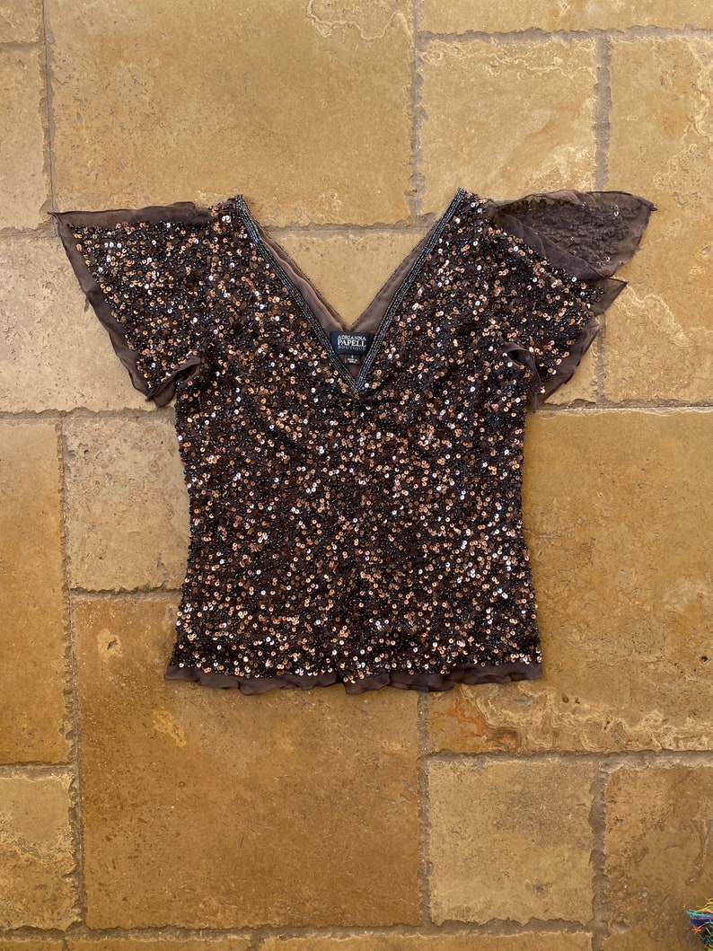 Puede incluir: Una blusa marr&oacute;n de lentejuelas con mangas cortas con volantes y cuello en V. La blusa tiene una etiqueta que dice "Adrianna Papell".