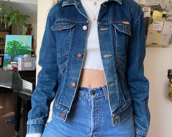 May include: A blue denim jacket with a button-up front and long sleeves. The jacket has a faded look and features copper-colored buttons.