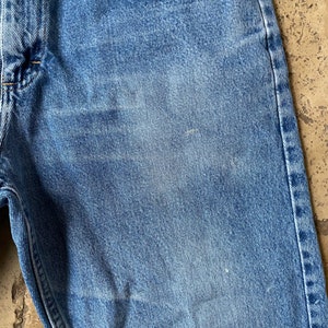 K&ouml;nnte beinhalten: Nahaufnahme einer blauen Jeanshose. Der Stoff hat einen verblassten, abgenutzten Look mit einer subtilen Textur. Die Jeans hat einen klassischen F&uuml;nftaschen-Stil.