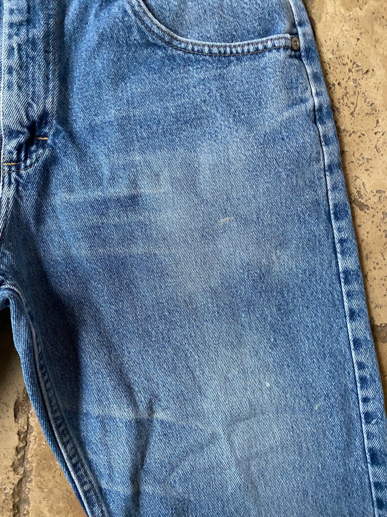 K&ouml;nnte beinhalten: Nahaufnahme einer blauen Jeanshose. Der Stoff hat einen verblassten, abgenutzten Look mit einer subtilen Textur. Die Jeans hat einen klassischen F&uuml;nftaschen-Stil.