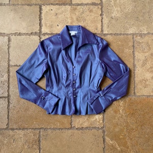 Puede incluir: Una blusa de color azul-violeta con un brillo lustroso. La parte superior de manga larga presenta un cuello, una botonadura frontal y una cintura plisada. Los puños tienen cierres de botón. La blusa está colocada sobre una superficie de baldosas de piedra.