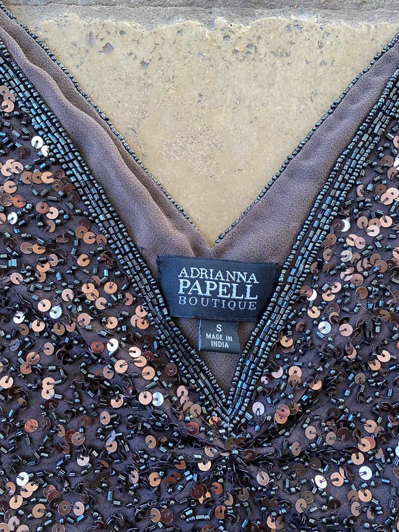 Puede incluir: Una blusa gris oscuro con lentejuelas y escote en V. La etiqueta dice "Adrianna Papell Boutique" y "S Made in India".