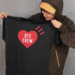 Op de afbeelding: Een zwart t-shirt met korte mouwen en een grote rode hartafbeelding op de borst. Binnenin het hart staat de witte tekst BTS CREW. Drie korte rode diagonale lijnen bevinden zich boven de rechterkant van het hartmotief op het t-shirt.